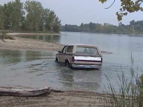 1995 Ford Bronco - Rear view of a brown and tan two-tone 1980s full-size Ford Bronco wading through shallow water off a sandy shoreline, with wheels submerged…