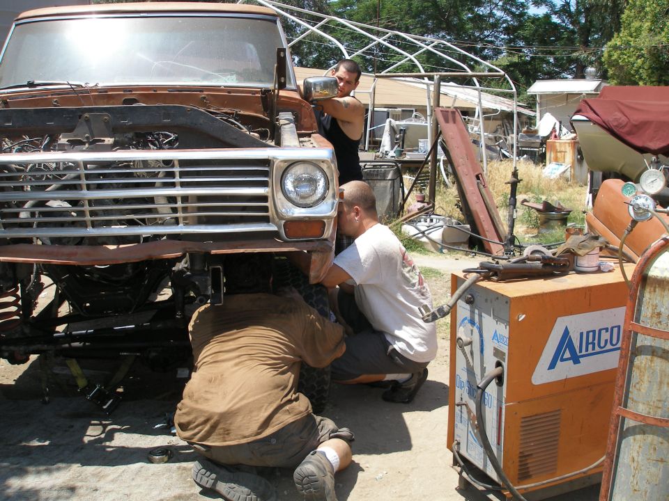 Three guys working on the front end of a rusty truck, with a welding machine nearby