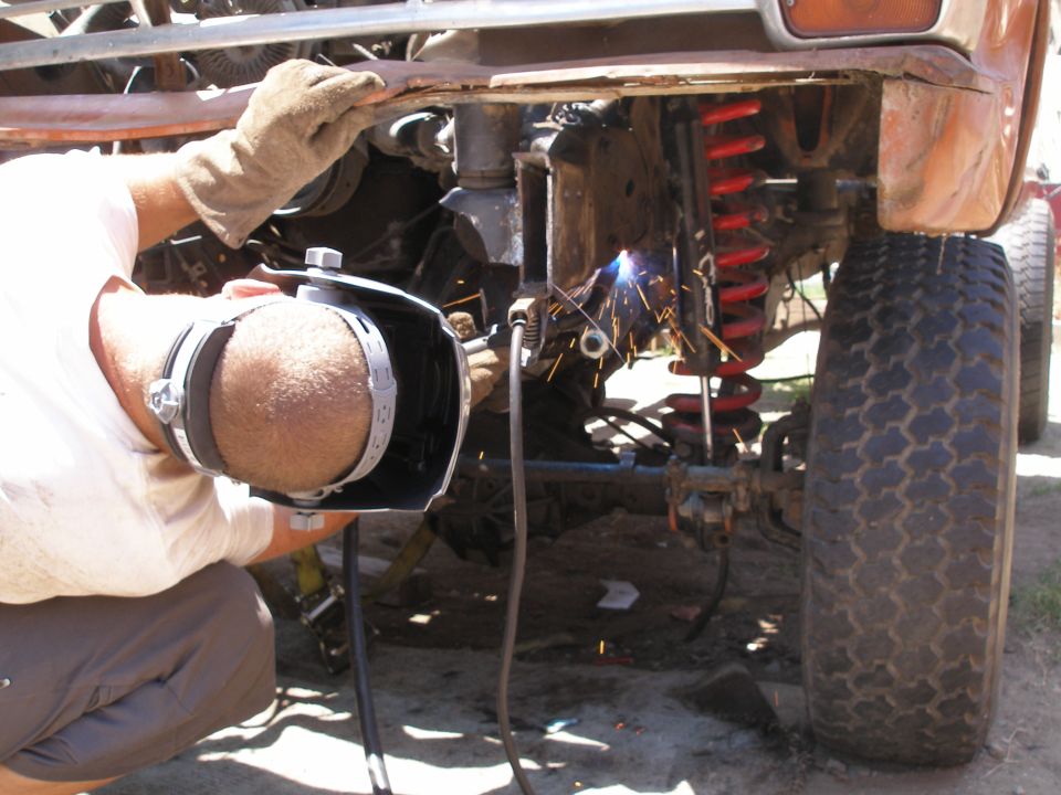 Man in welding mask works on rusty vehicle frame near red coil spring and large tire