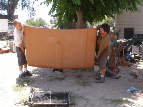 1969 Ford F-250 - Two men carry a large truck hood, possibly for a restoration project, in a cluttered outdoor area.