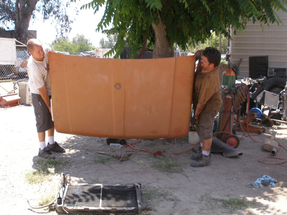 Two guys carrying a large, orange truck hood outdoors. A radiator is on the ground