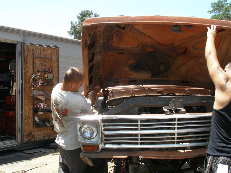 Orange truck front with hood open, men working on engine. Grille suggests early 1970s