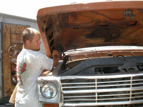 1969 Ford F-250 - A man works on the engine of a rusty Ford truck. The grille and headlight are visible.