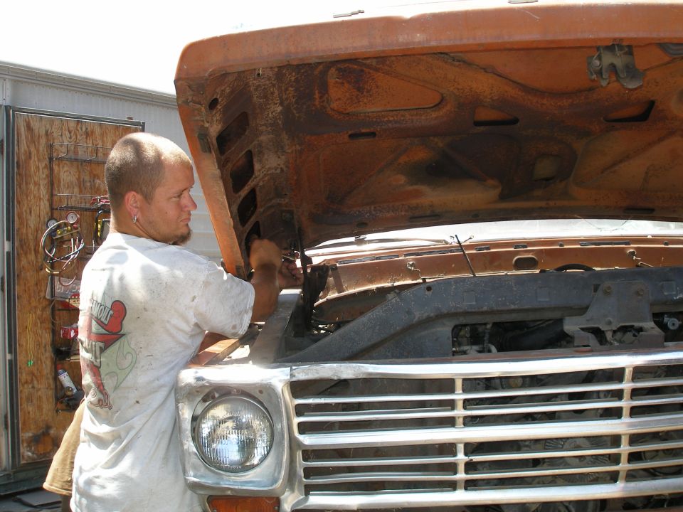 Man working on a rusty 1973 Ford F-Series truck under the hood. The grille is visible