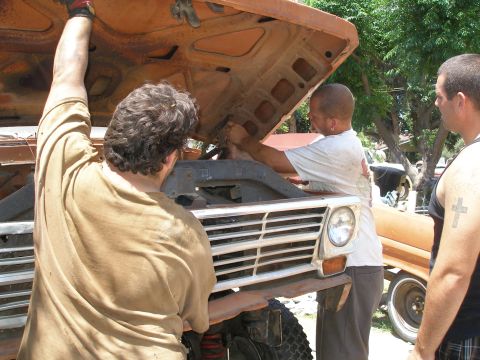 1969 Ford F-250 - Three men are gathered around the front of an orange truck with its hood open, appearing to work on the engine.