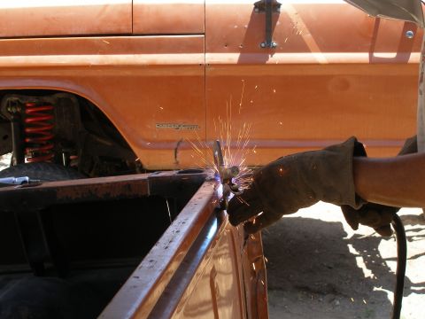 1969 Ford F-250 - A person in gloves welds a metal frame on the side of an orange truck bed, with sparks flying.