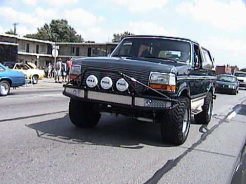 1995 Ford Bronco - Front view of a lifted dark green 1990s full-size Ford Bronco on a street at a cruise-in, fitted with a black tube brush guard carrying thr…