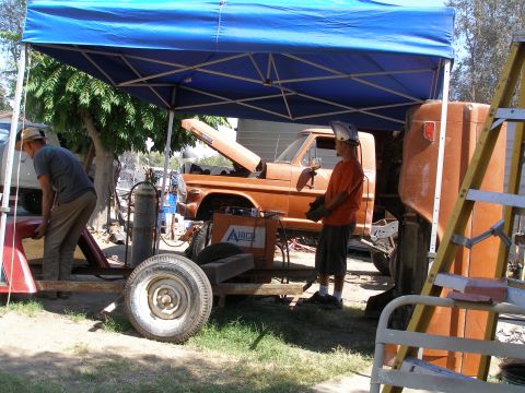 1969 Ford F-250 - A man in an orange shirt and welding helmet stands by an orange pickup truck with its hood up, undergoing work outdoors.