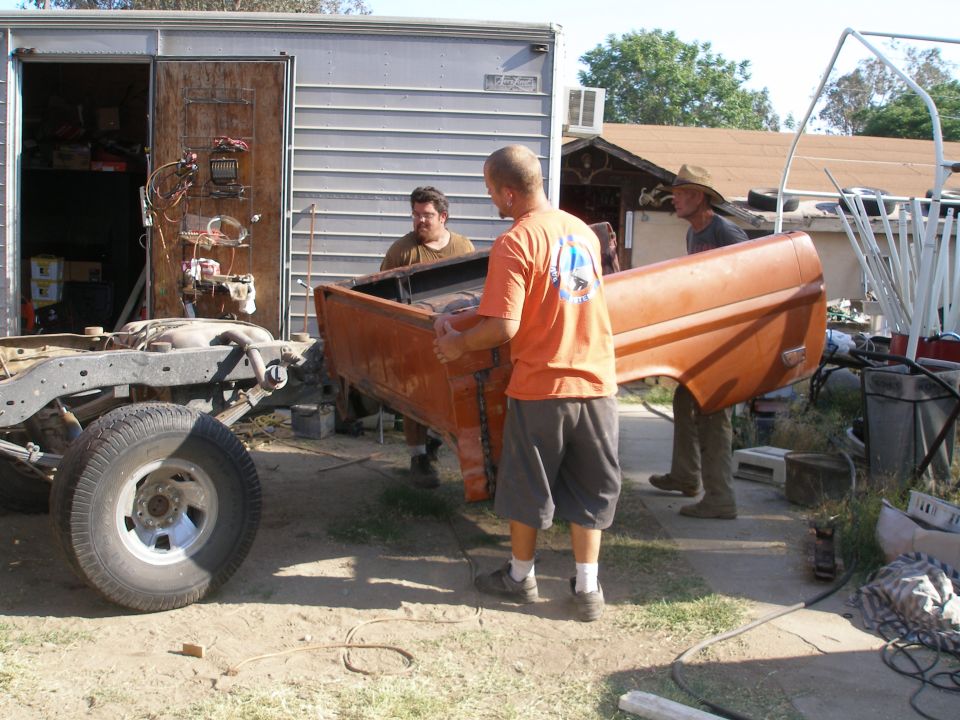 Three men carrying an orange truck bed near a bare chassis and a trailer