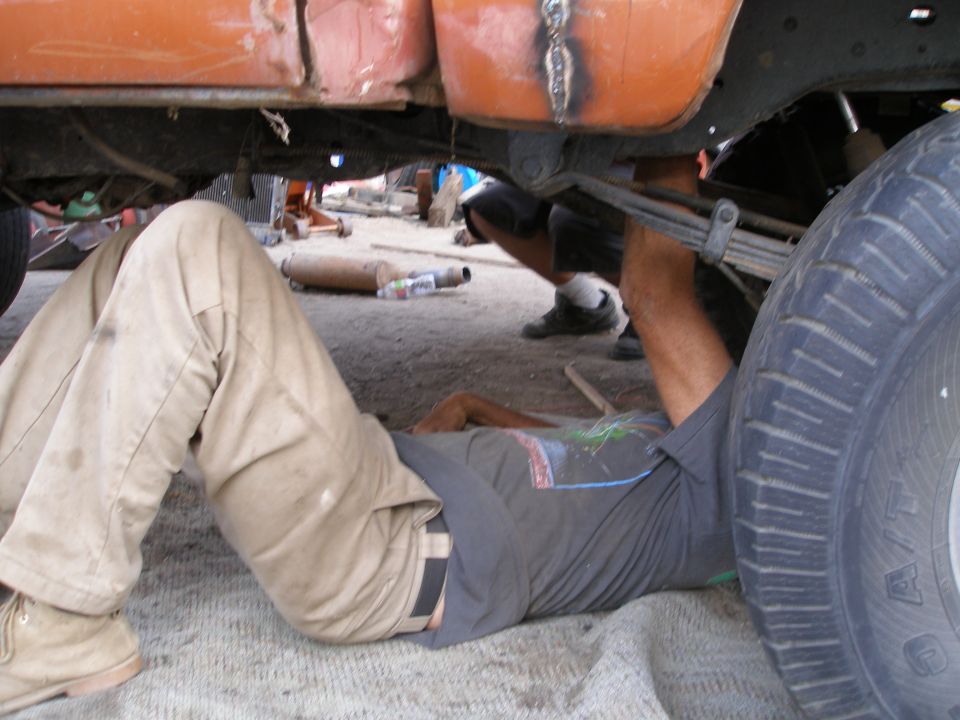 Guy working under an orange truck, near the rear tire and leaf springs. Another person is visible in the background