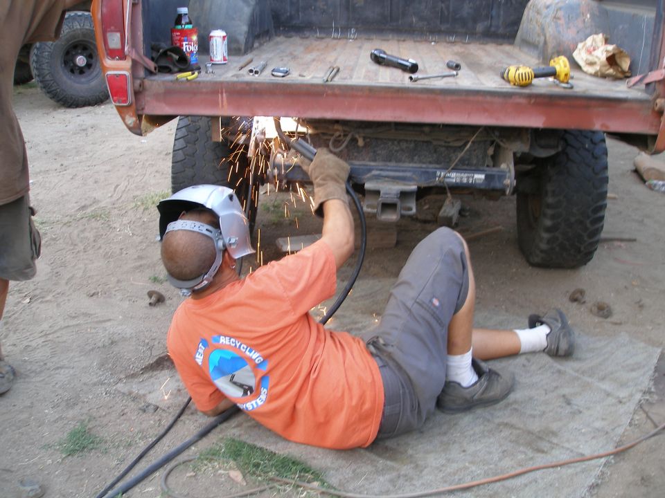 Welding on the rear frame of a red pickup truck bed, with another vehicle visible to the left