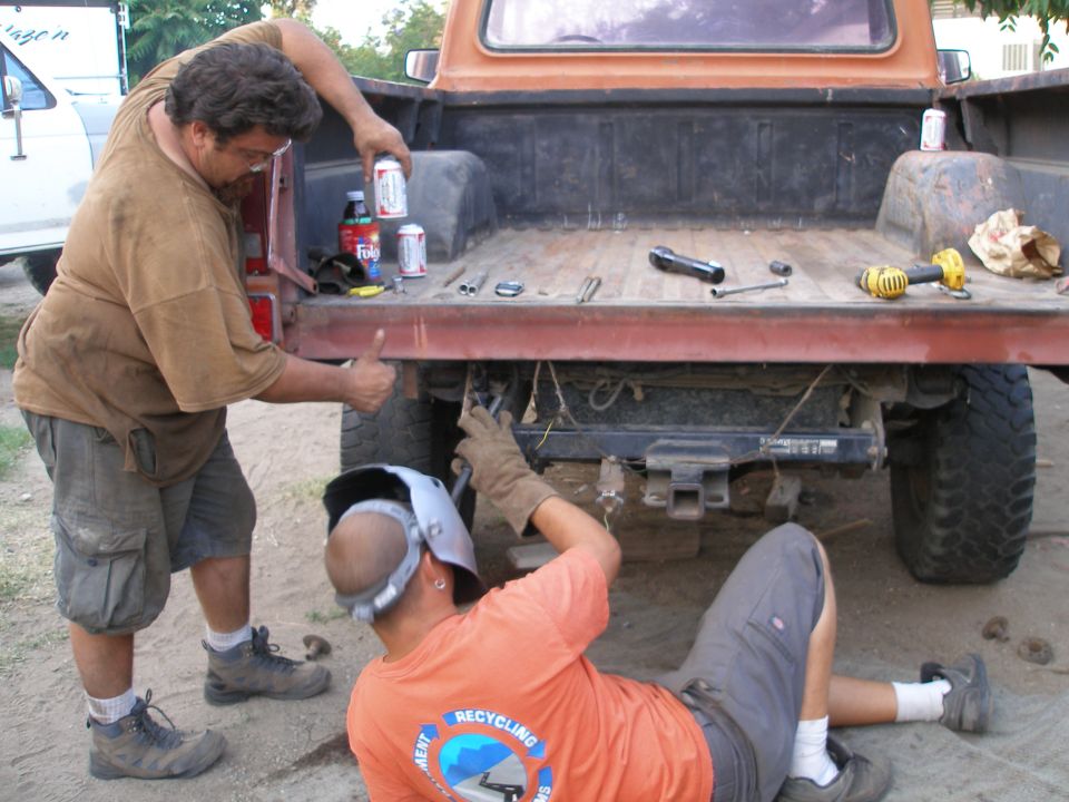 Two guys are working on the back of an orange pickup truck. One is standing and holding a can, the other is lying down with a welding mask