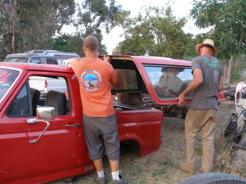 1969 Ford F-250 - Two men work on removing a camper shell from a red pickup truck in an outdoor setting with other vehicles present.