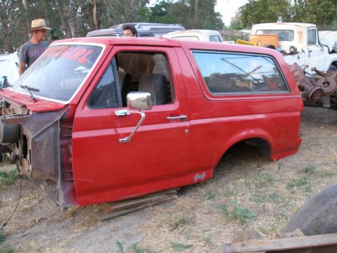 1969 Ford F-250 - This red Ford Bronco body, missing its front clip and rear axle, sits on wood planks in a yard with other vehicles.