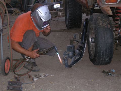 1969 Ford F-250 - A person in a welding mask uses a torch on a vehicle's suspension component, with a large tire visible nearby.