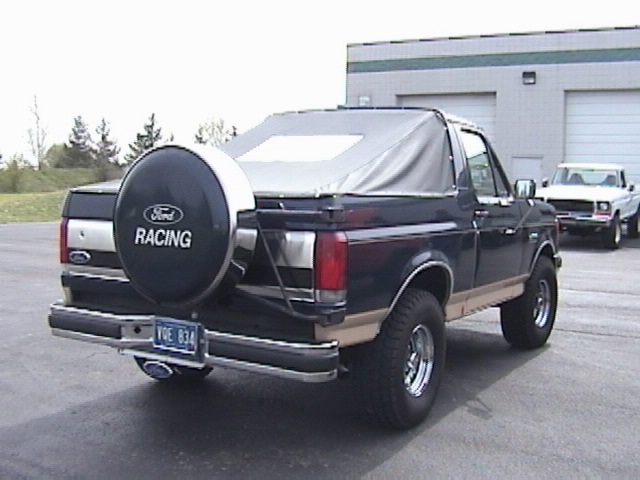 1995 Ford Bronco rear view with soft top and spare tire carrier, another white truck in background