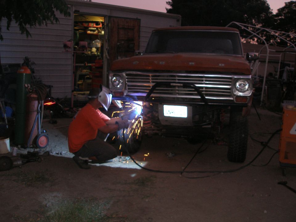Orange Ford truck with a custom bumper, being welded by a person in a mask