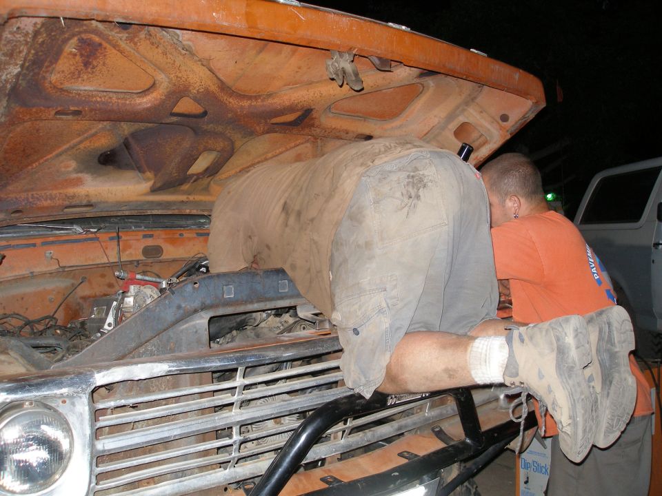Two people working on the engine of an orange truck, another vehicle visible to the right