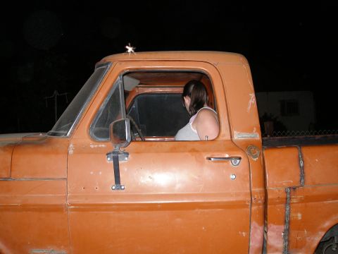 1969 Ford F-250 - A woman sits in the cab of an orange pickup truck at night, with visible repairs on the truck bed.