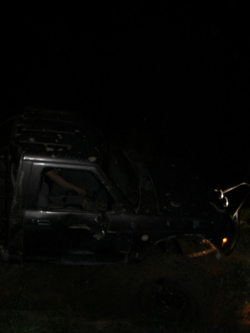 Dark SUV with headlights on, off-road at night. Driver's arm visible through window