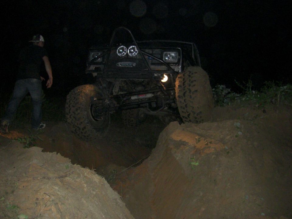 Off-road vehicle with large tires navigating a dirt obstacle at night. A person stands to the left