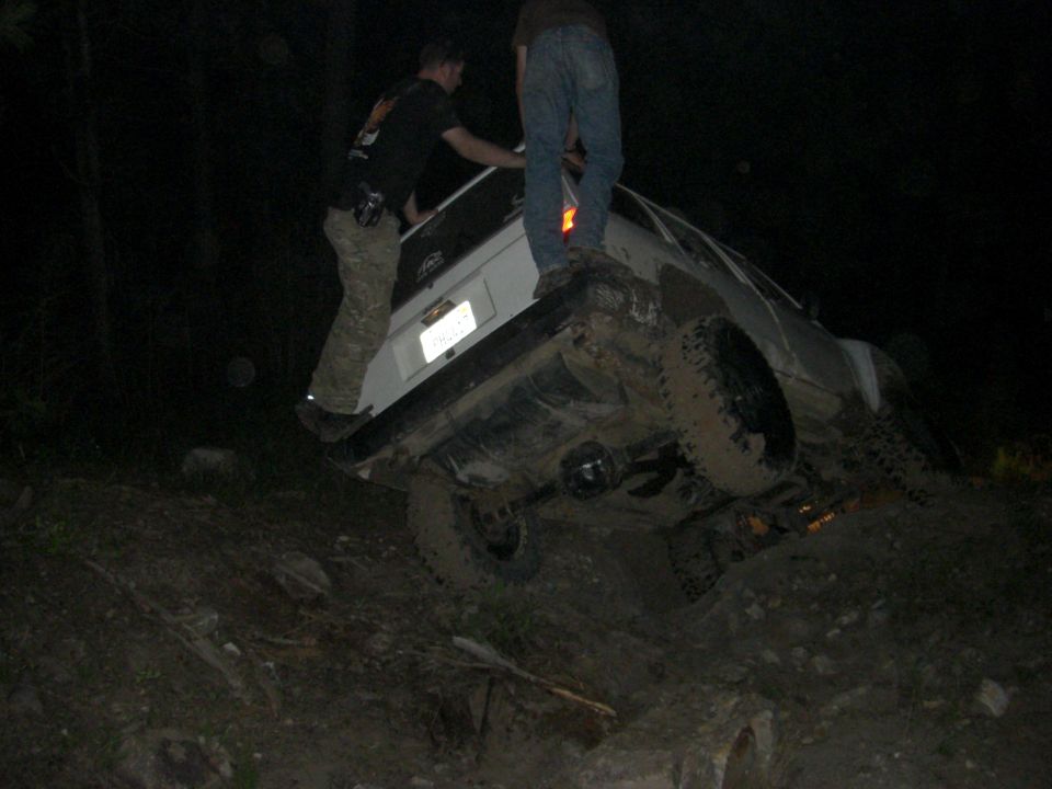 White SUV tilted on a muddy hill at night with two people standing on it