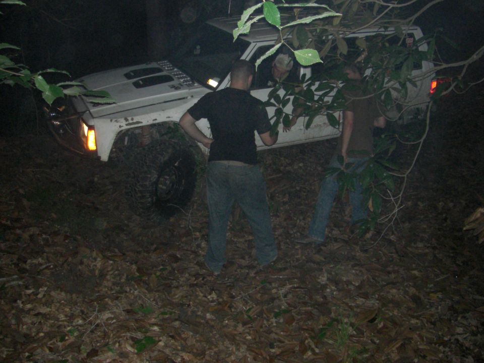 White SUV stuck in woods at night with people around it