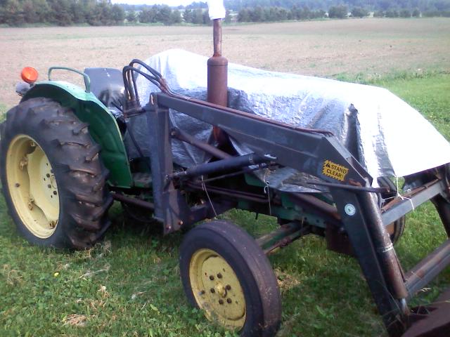 Green and yellow farm tractor with front loader and silver tarp parked in grass by a field