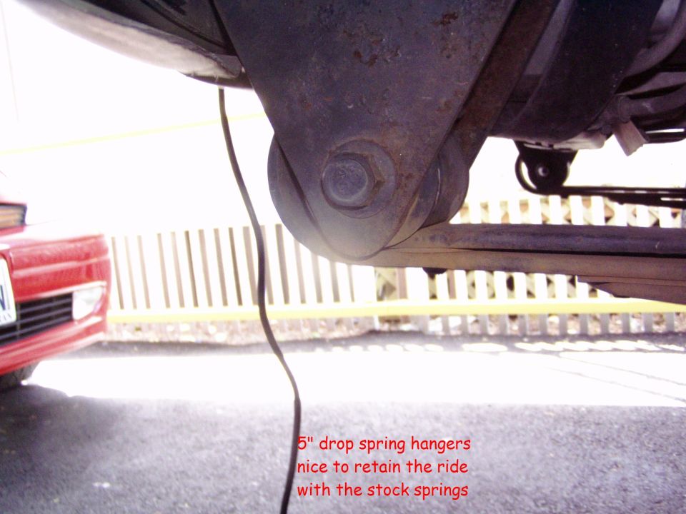 close-up of a vehicle's rear suspension with leaf springs and a drop spring hanger; red vehicle visible on left
