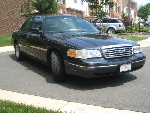 1999 Ford Crown Victoria - A dark sedan with a chrome grille is parked on a street, with a silver SUV in the background.