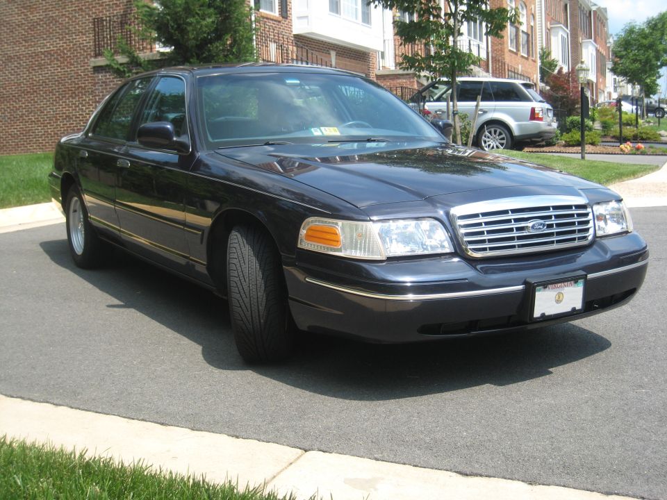Dark sedan parked on asphalt, front 3/4 view. Chrome grille, clear headlights, chrome trim. Another SUV is visible in the background