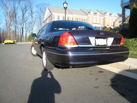 1999 Ford Crown Victoria - Rear view of a dark blue 1999 Ford Crown Victoria parked on a street, with a yellow car visible in the distance.