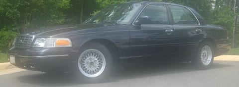 1999 Ford Crown Victoria - A dark sedan is parked on a paved surface with trees in the background.