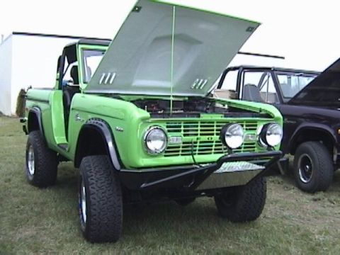 1995 Ford Bronco - A bright green first-generation Ford Bronco is displayed with its hood open, revealing the engine bay. A second Bronco is visible to the ri…