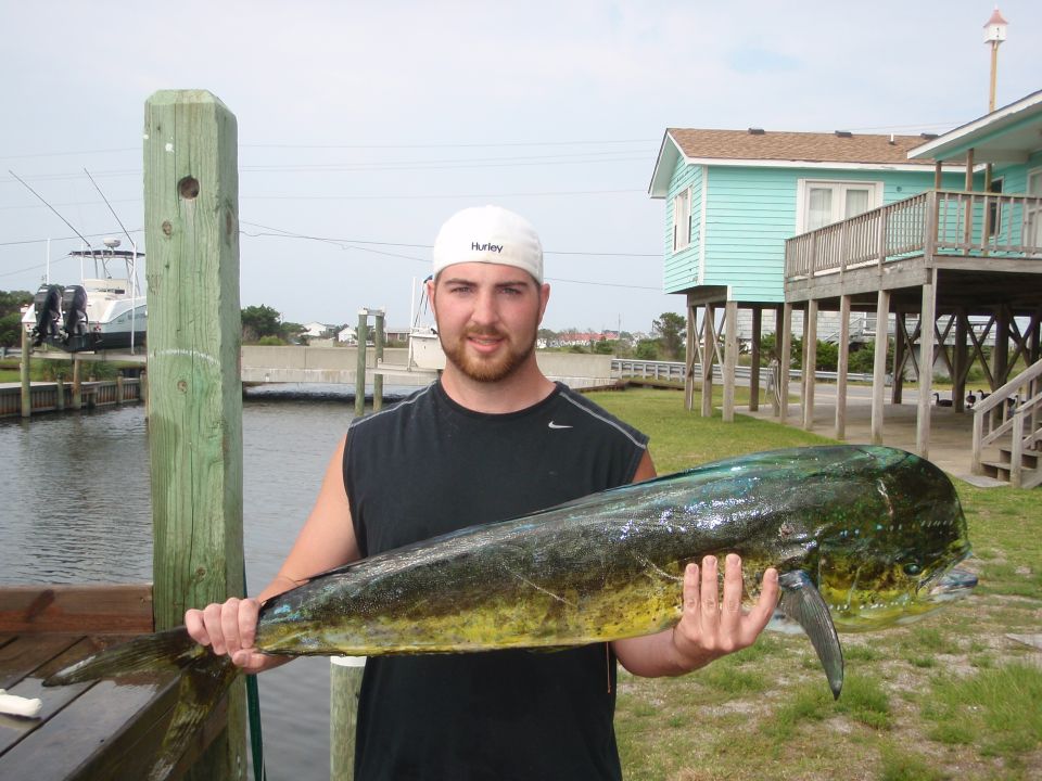 Man on a dock holding a dolphin fish, with a light blue house on stilts in the background