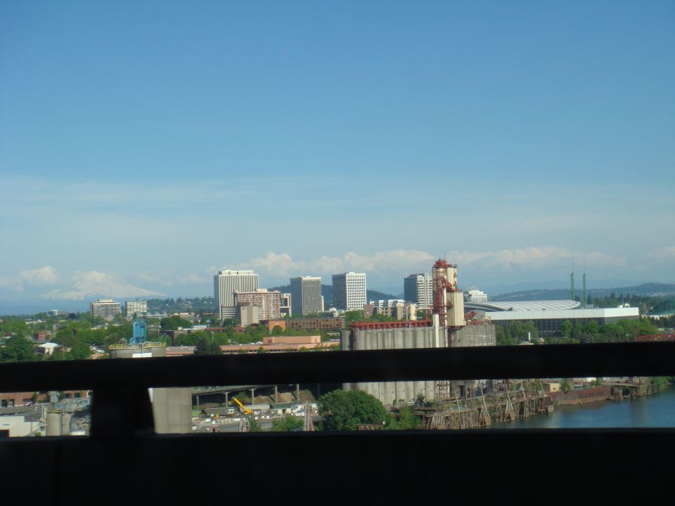 City skyline with industrial structures and a river, mountains visible on the horizon under a blue sky