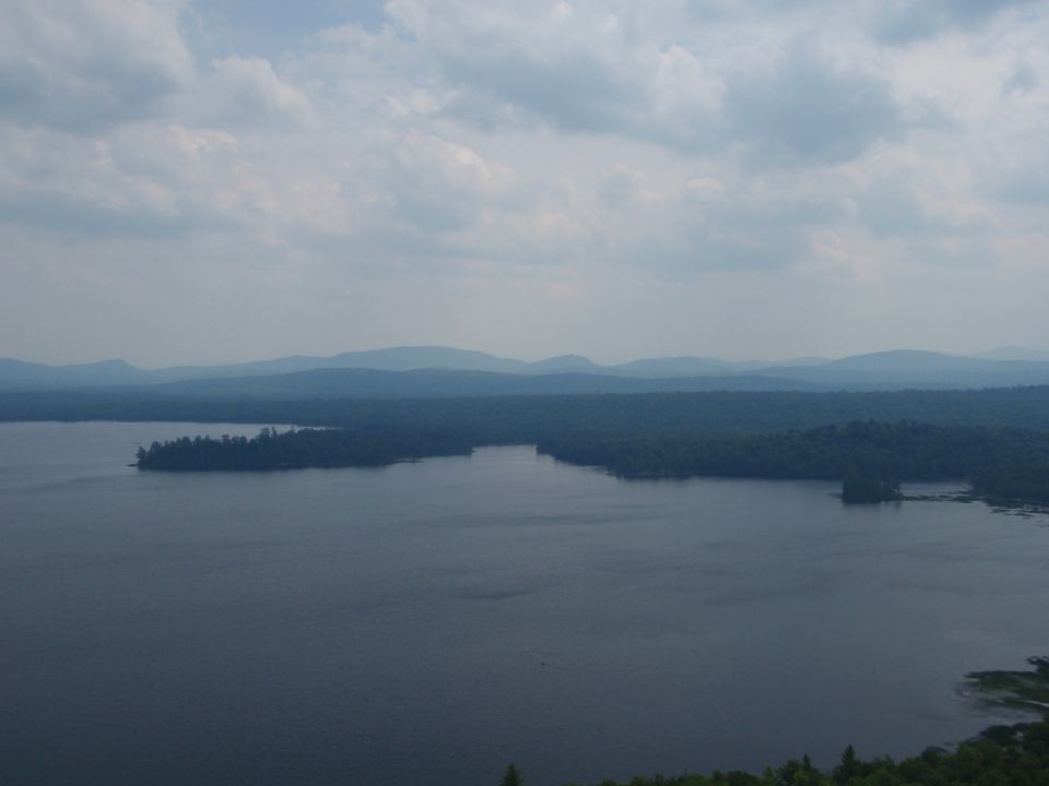 Wide view of a large lake with tree-covered islands and distant blue mountains under a cloudy sky. Watermark at bottom right