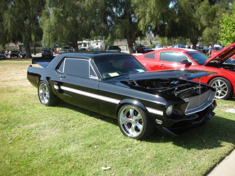 1992 Ford Crown Victoria - A black 1967 Ford Mustang fastback with a white stripe and custom wheels is displayed at a car show, with its hood open.
