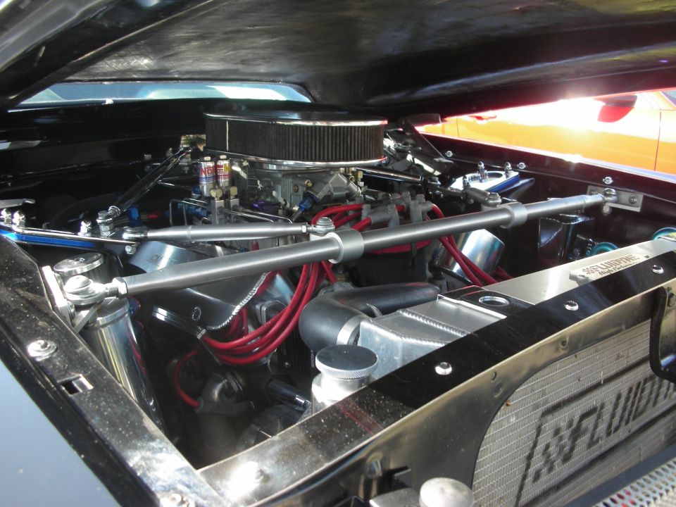 engine bay with V8, chrome parts, red spark plug wires, and a large air filter. Another car is visible in the background