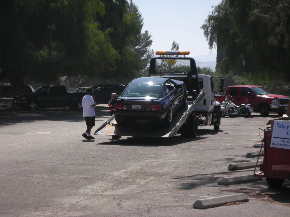 Dark blue coupe being loaded onto a flatbed tow truck, with a red pickup truck and motorcycle parked nearby