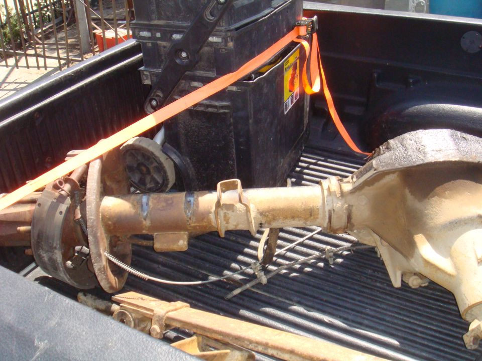 Rear axle assembly with drum brakes and brake lines secured in a pickup truck bed with an orange strap and a toolbox