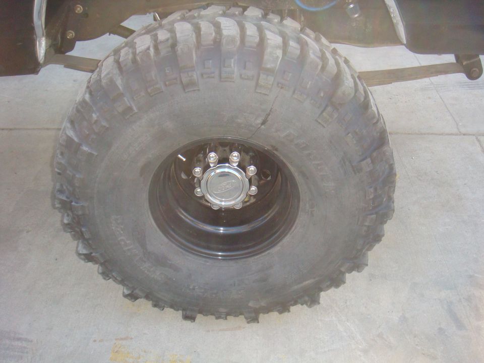 Close-up of a large, knobby off-road tire on a black rim with a chrome center cap. Part of the vehicle's suspension is visible above