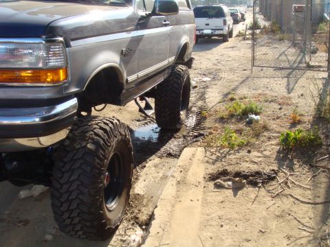 1992 Ford Bronco - A lifted 1992 Ford Bronco with oversized tires is shown off-road, with other vehicles in the background.