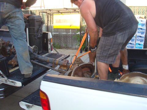 1992 Ford Bronco - Workers are carefully maneuvering a large vehicle axle into the bed of a pickup truck.