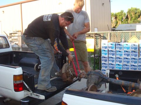 1992 Ford Bronco - Two men use straps to load heavy axles into the beds of two pickup trucks.