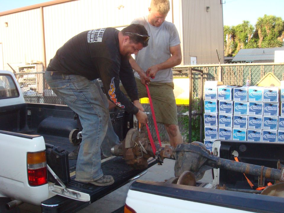 Truck beds with axles being loaded, men with straps, stack of Bud Light cases