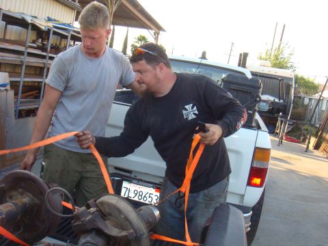 1992 Ford Bronco - Two men are seen securing parts in the bed of a white pickup truck using orange straps. The license plate is visible.