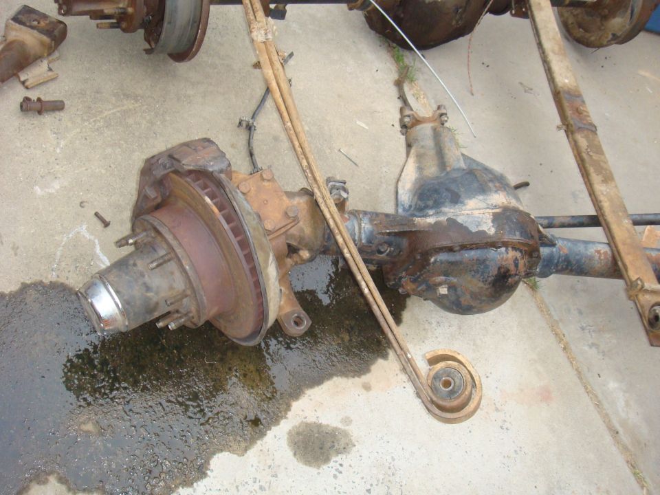 Vehicle axles and leaf springs disassembled on concrete. One axle has a brake rotor and hub assembly