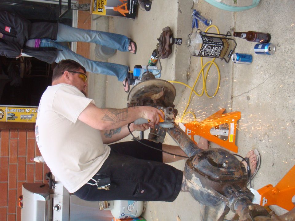 Guy grinding on a vehicle axle with sparks flying. Two people stand behind him