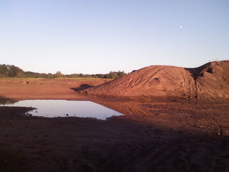 Red clay dirt mound and water-filled pit with tire ruts at sunset, no vehicle in frame